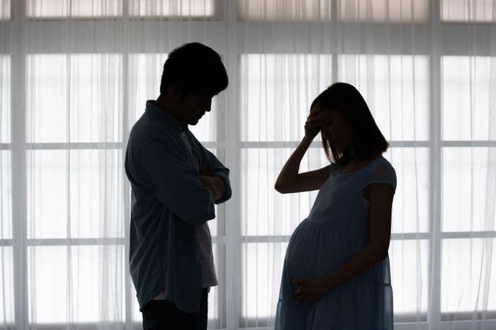Silhouettes of a man and pregnant woman standing apart, looking upset, in a room with closed curtains behind them