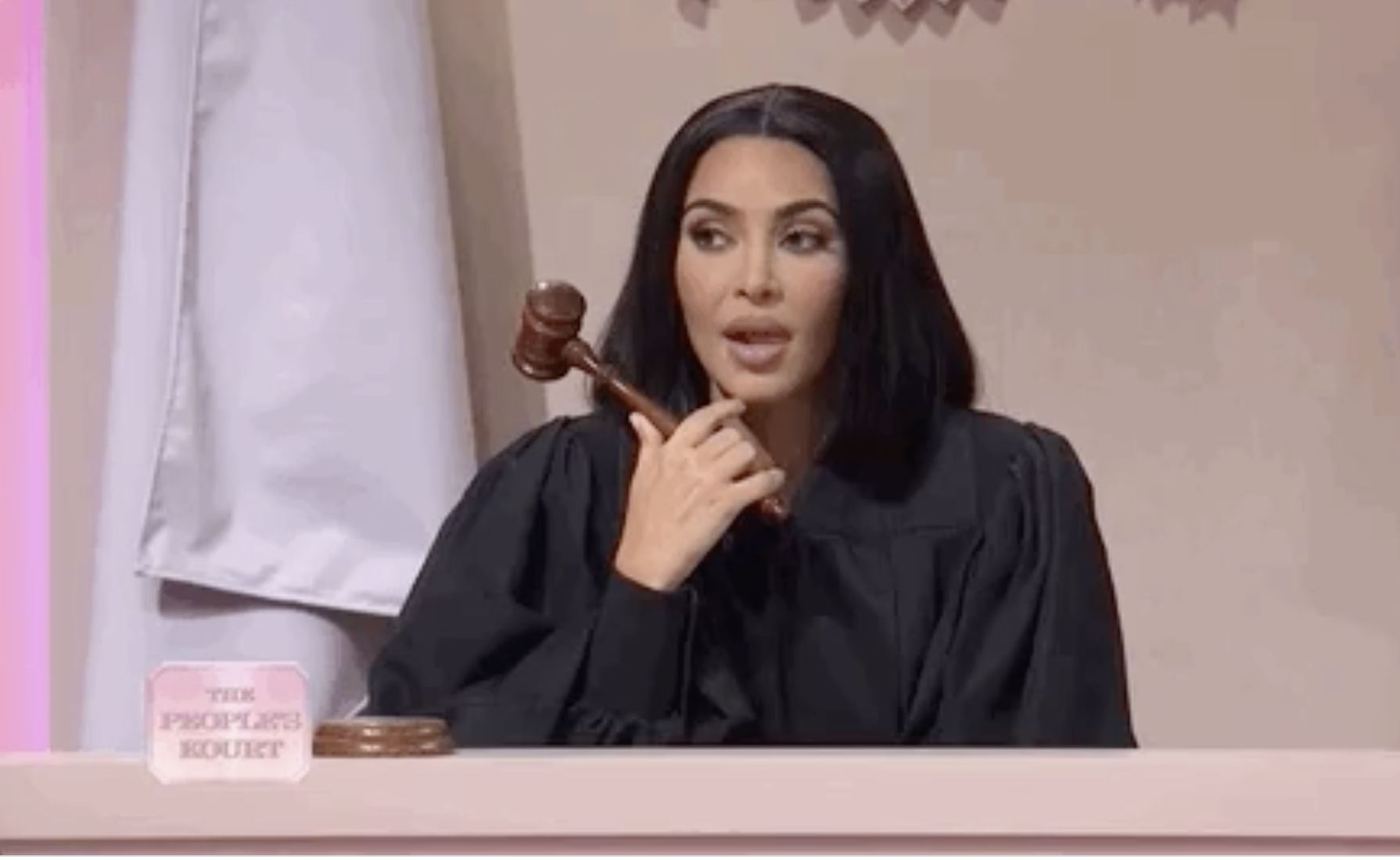 Kim Kardashian wearing a judge&#x27;s robe, seated behind a bench with a gavel in hand, looking thoughtful