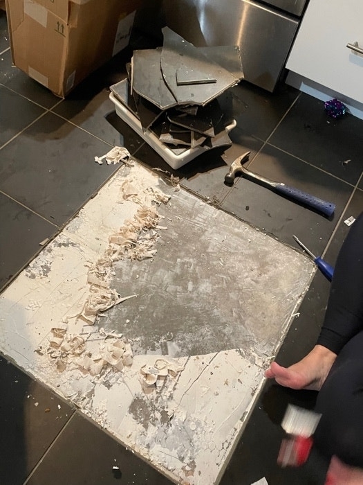 Damaged kitchen floor with debris and a person holding a hammer, indicating renovation or repair work in progress