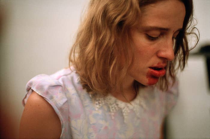 Woman in patterned dress and blood on her mouth looks down with an emotional expression