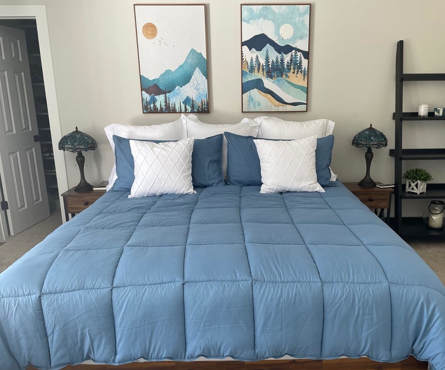 A neatly made bed with a blue comforter, white and blue pillows, flanked by two nightstands and decorative lamps, with wall art above