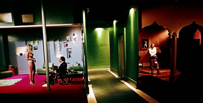 Scene with two characters in an interior room with green lighting and a pole, one seated in a wheelchair. Another character is in a room across the hall