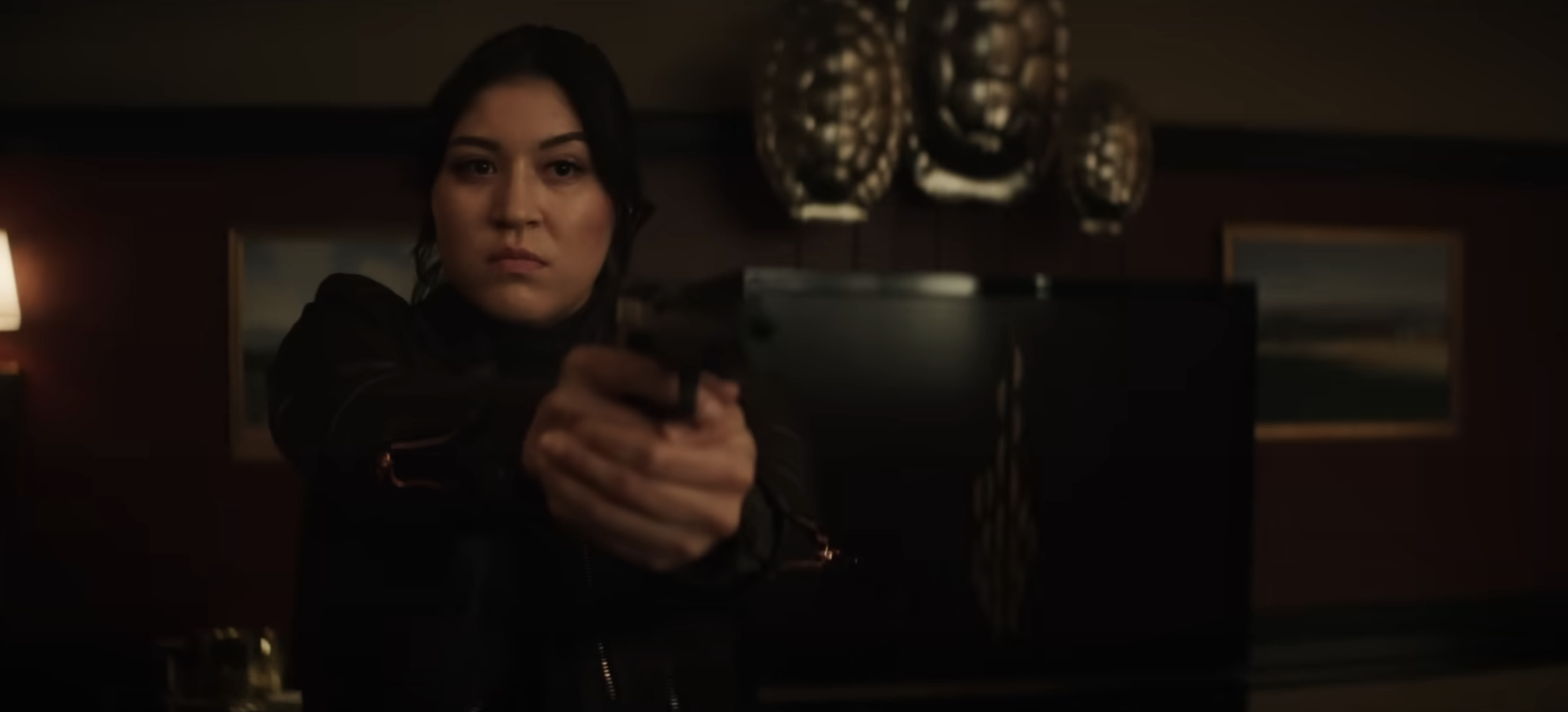 Maya aiming a gun with focus and determination in a dimly lit room