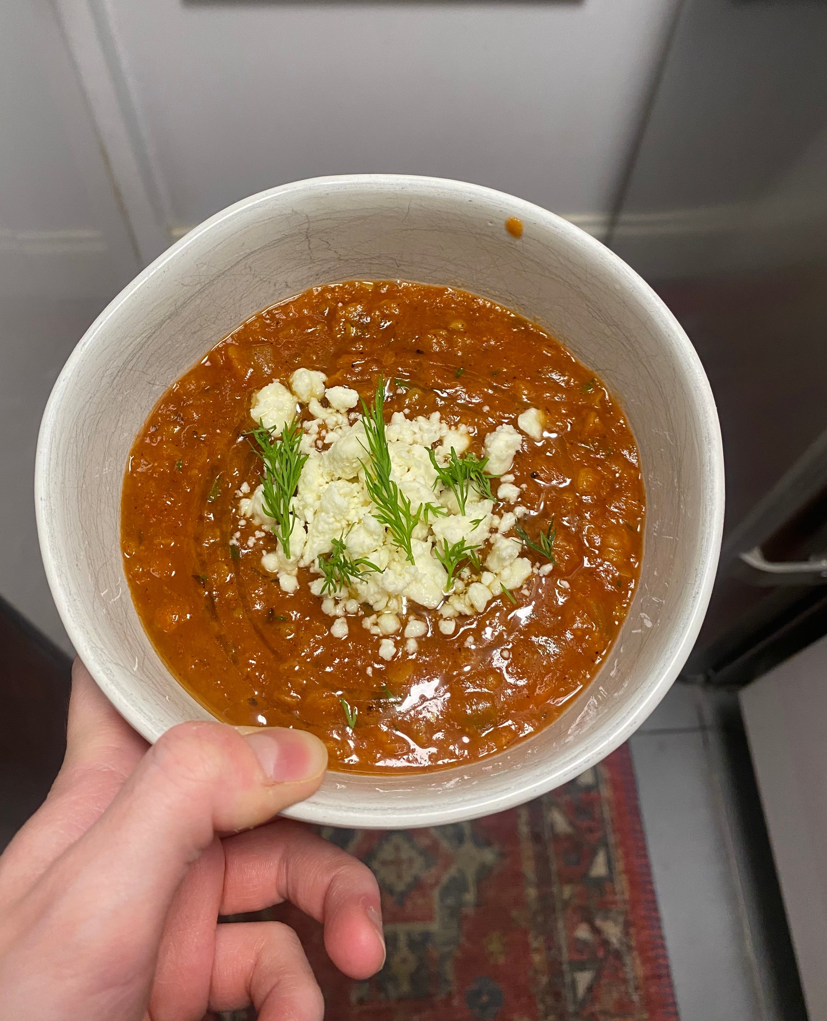 A person holding a bowl of tomato soup garnished with crumbled cheese and herbs