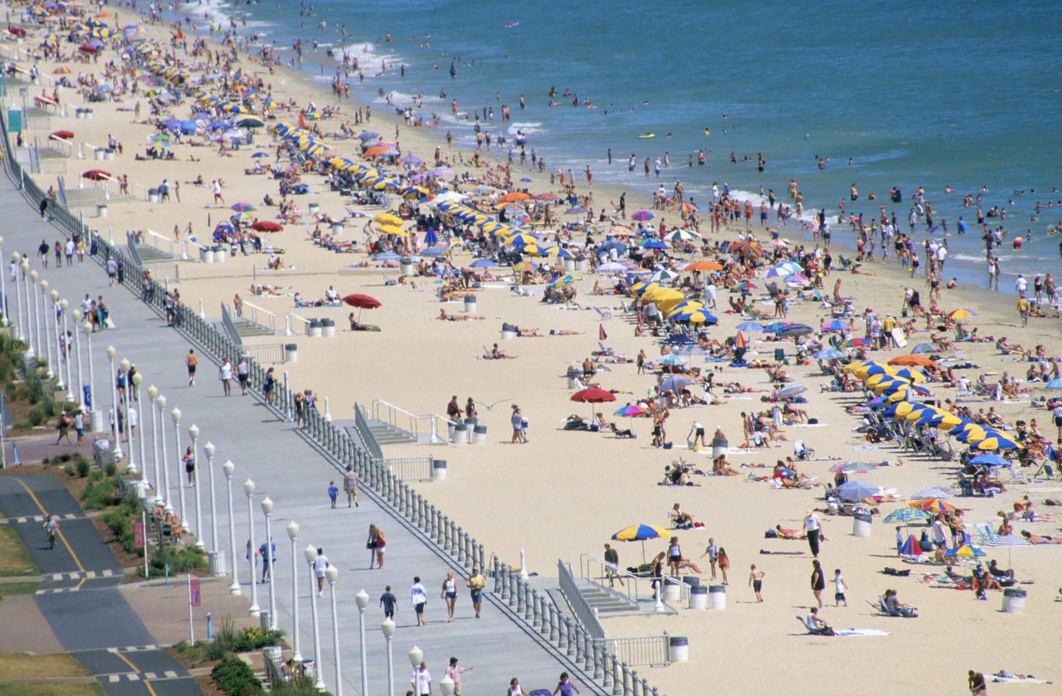 Crowded beach with people sunbathing and umbrellas, adjacent to a walkway