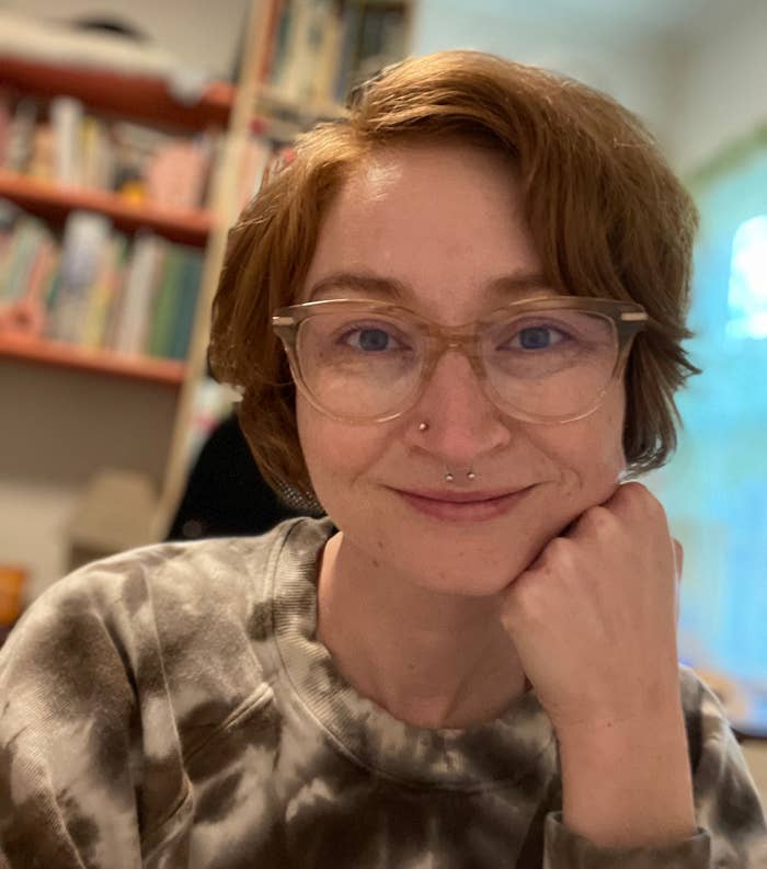 Woman smiling at camera with glasses and a septum piercing, resting her chin on her hand