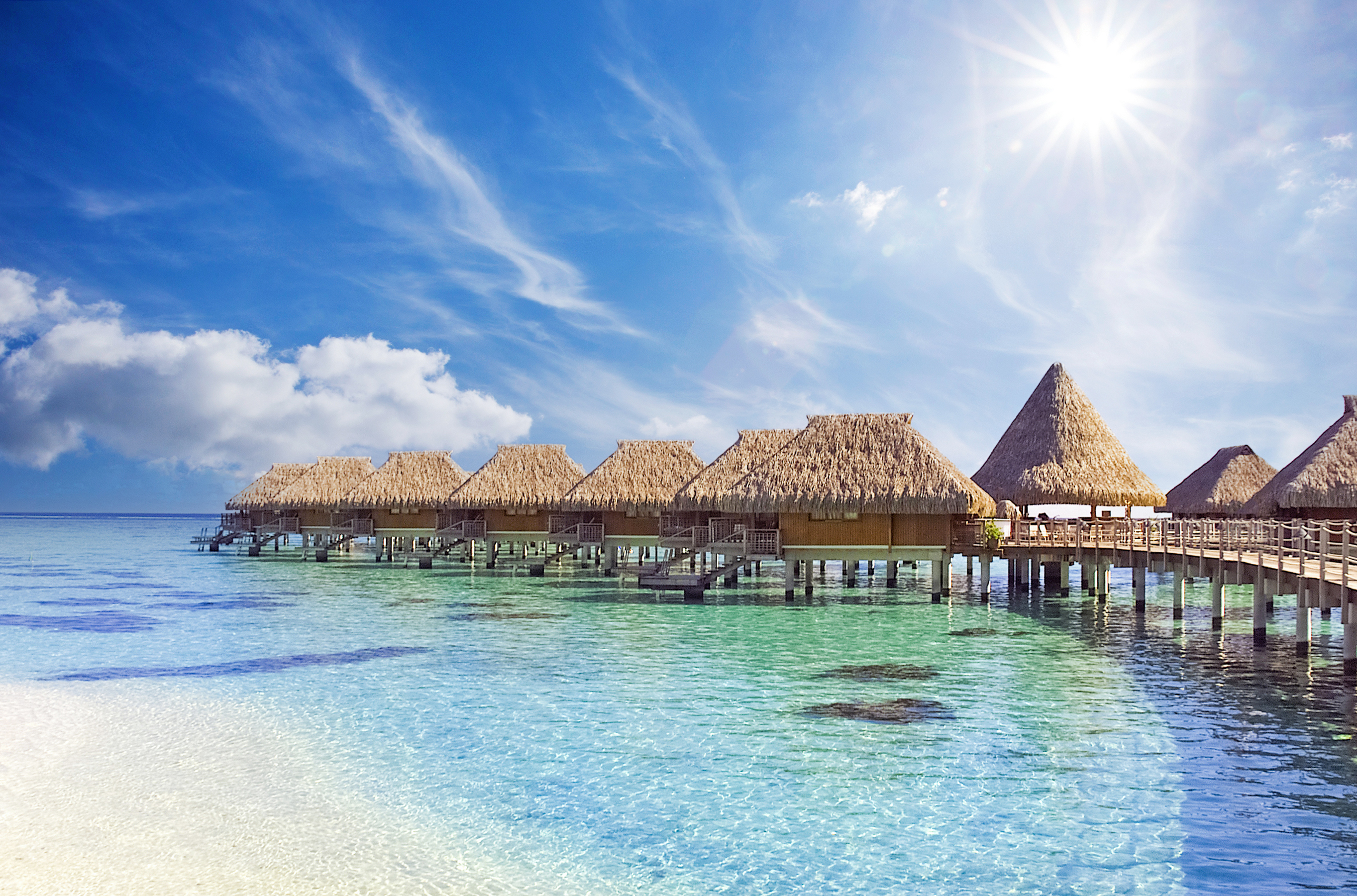 Overwater bungalows extend from a wooden pier into a calm sea under a sunny sky
