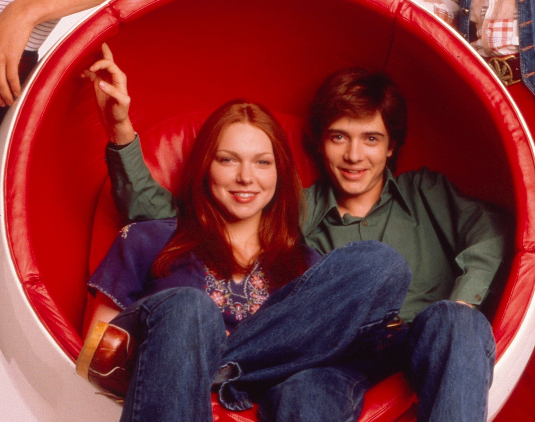 Donna Pinciotti and Eric Forman characters sitting in a large red bowl chair, in casual 70s attire, from &quot;That &#x27;70s Show.&quot;