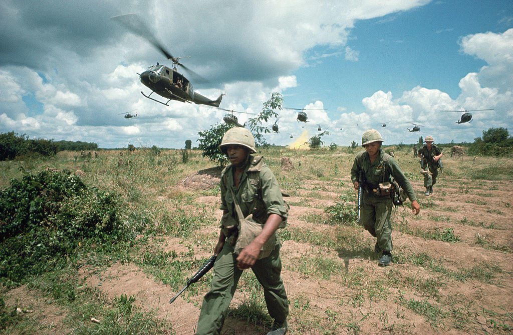 Soldiers walk in a field with helicopters flying overhead, embodying a historical military scene