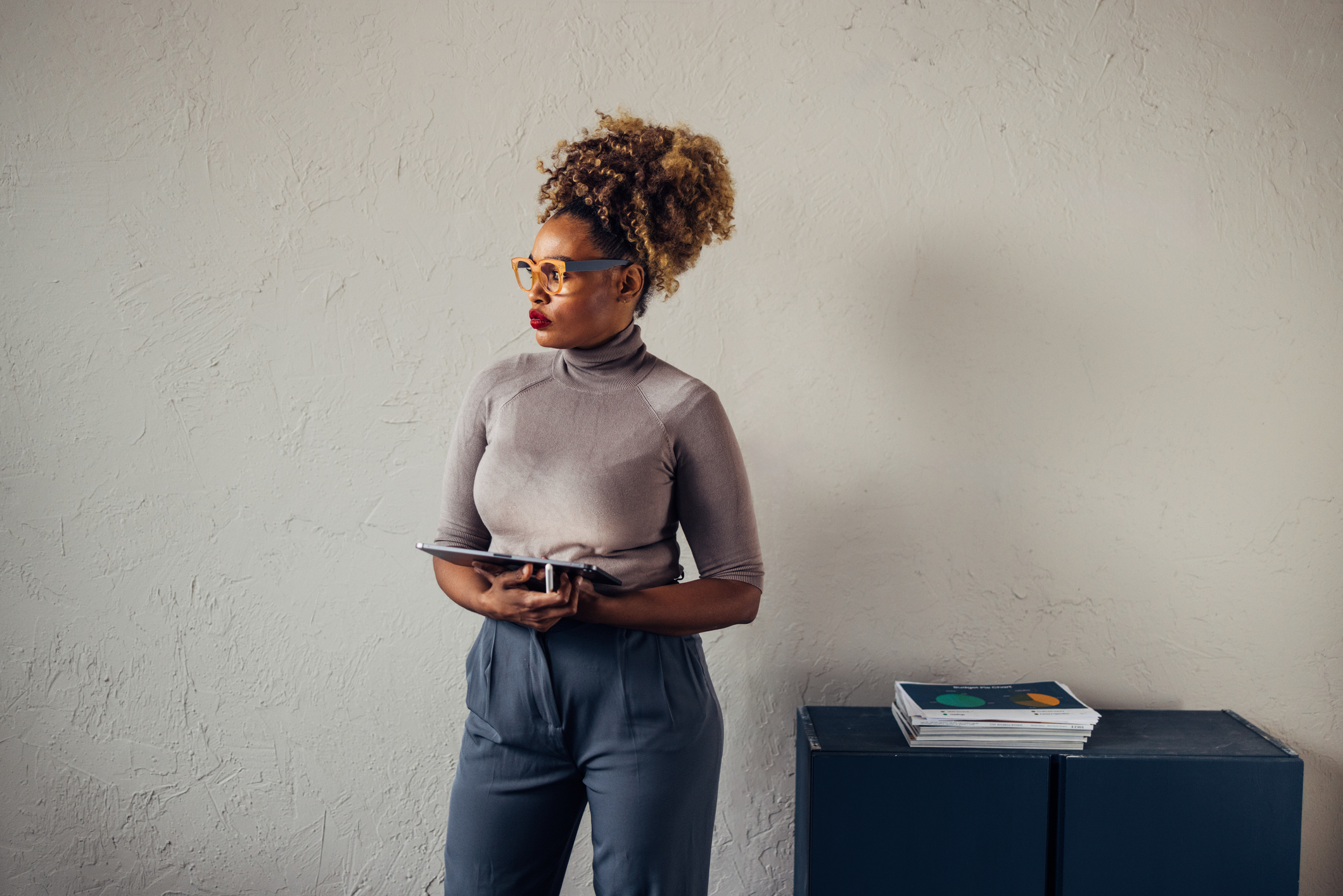 Woman in business casual attire with tablet looking to the side, standing indoors near a cabinet