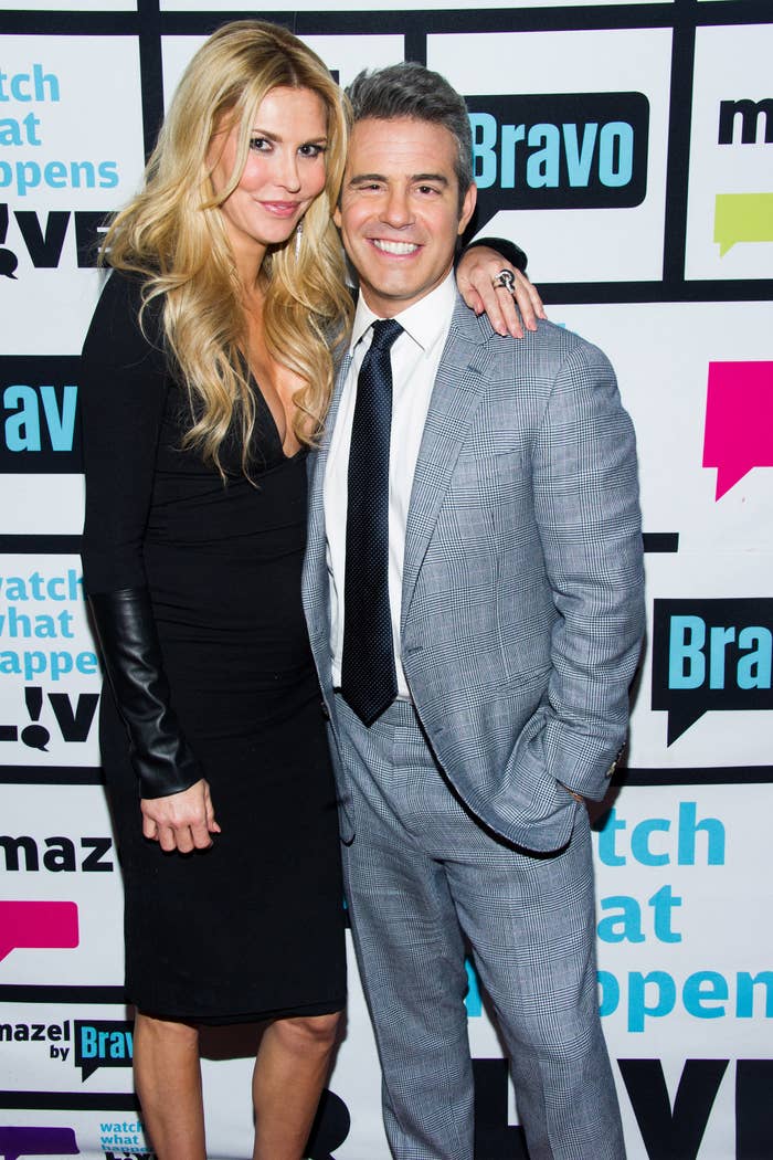 Brandi Glanville and Andy Cohen stand together at a media event, Brandi in a black dress, Andy in a gray suit