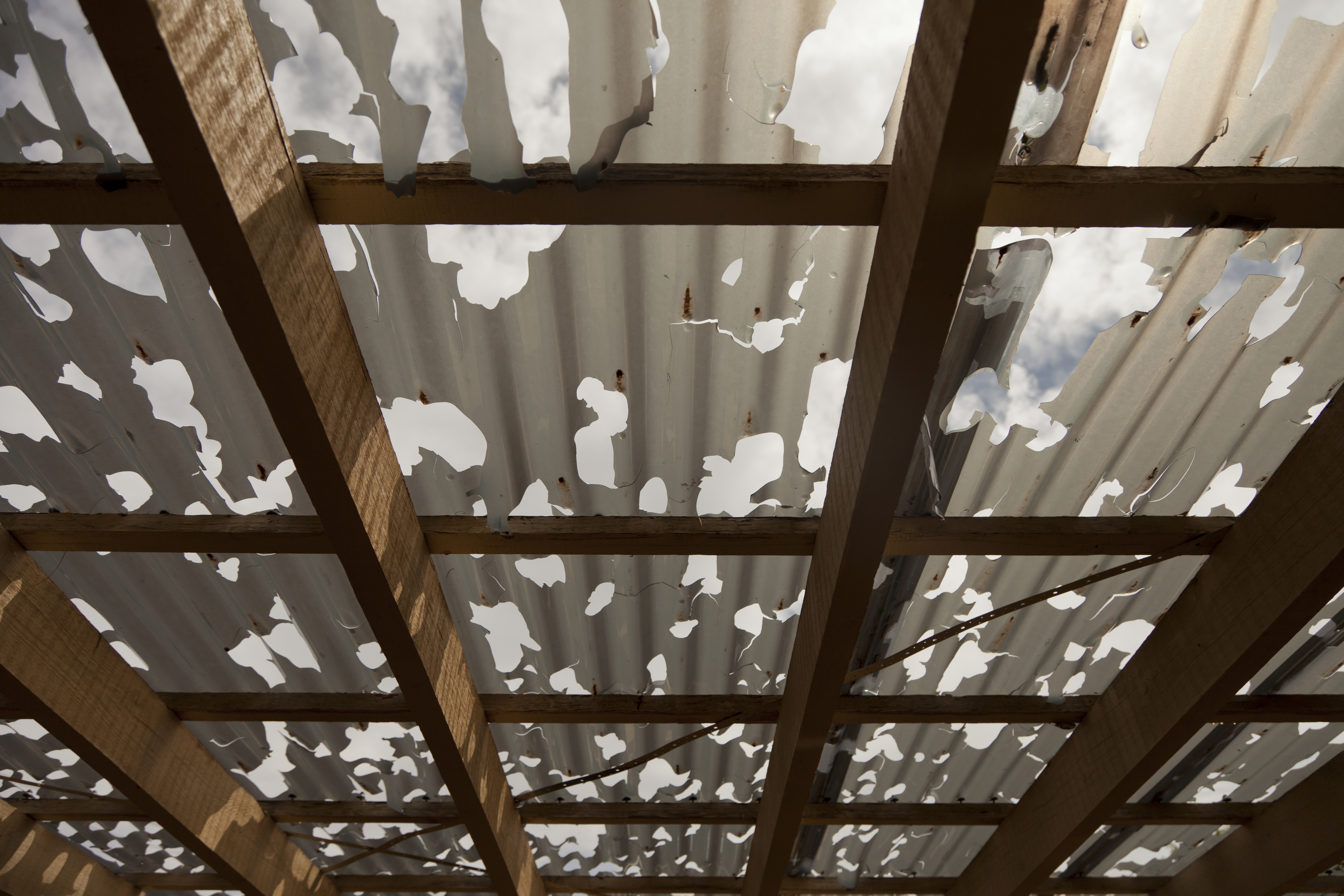 Damaged tin roof with holes, viewed from below, showing wooden beams against the sky