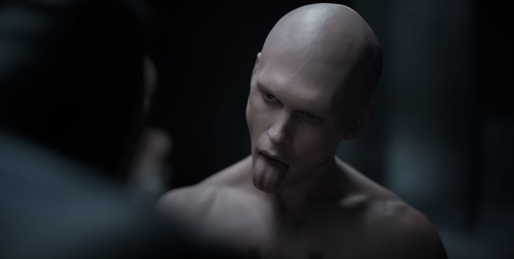 Austin, with a bald head, sticks his tongue out at someone in a dimly lit room