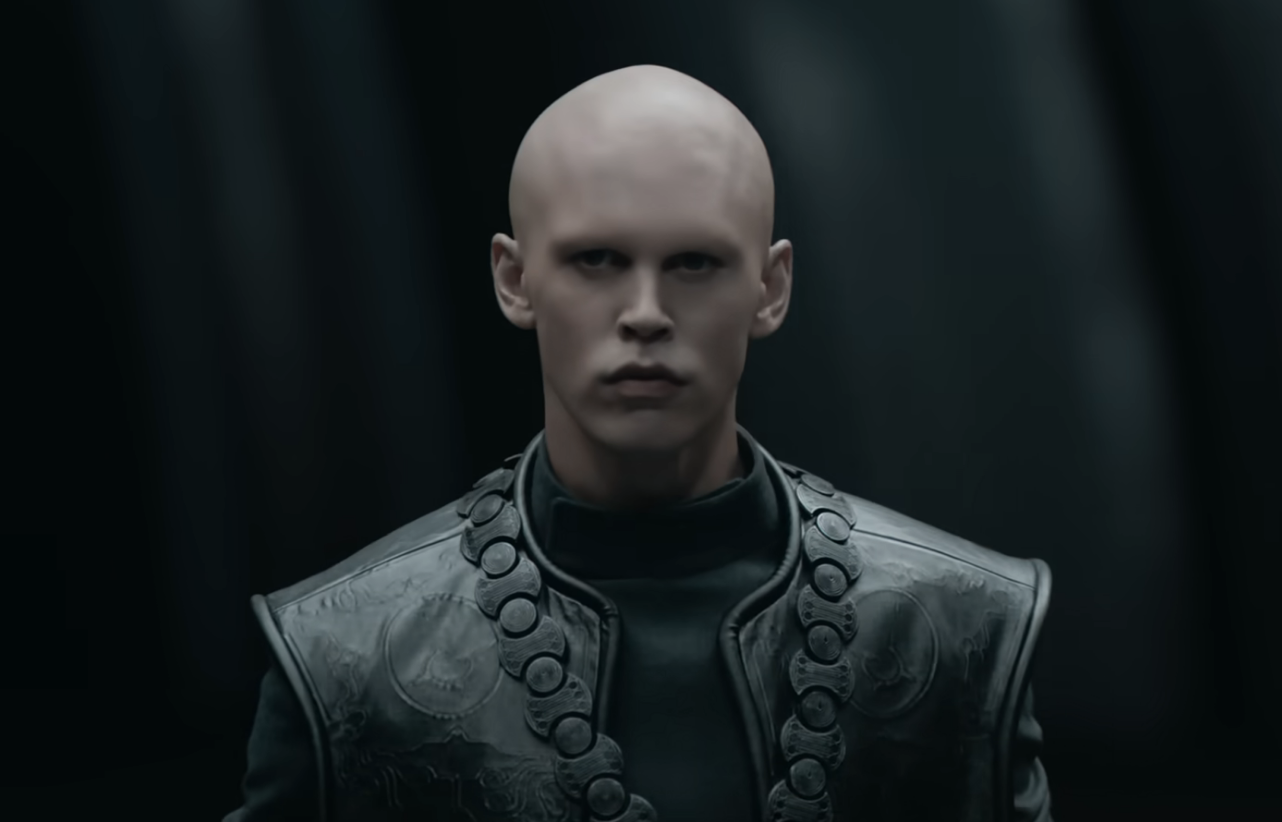 Austin with a bald head wearing a detailed leather jacket and looking solemn
