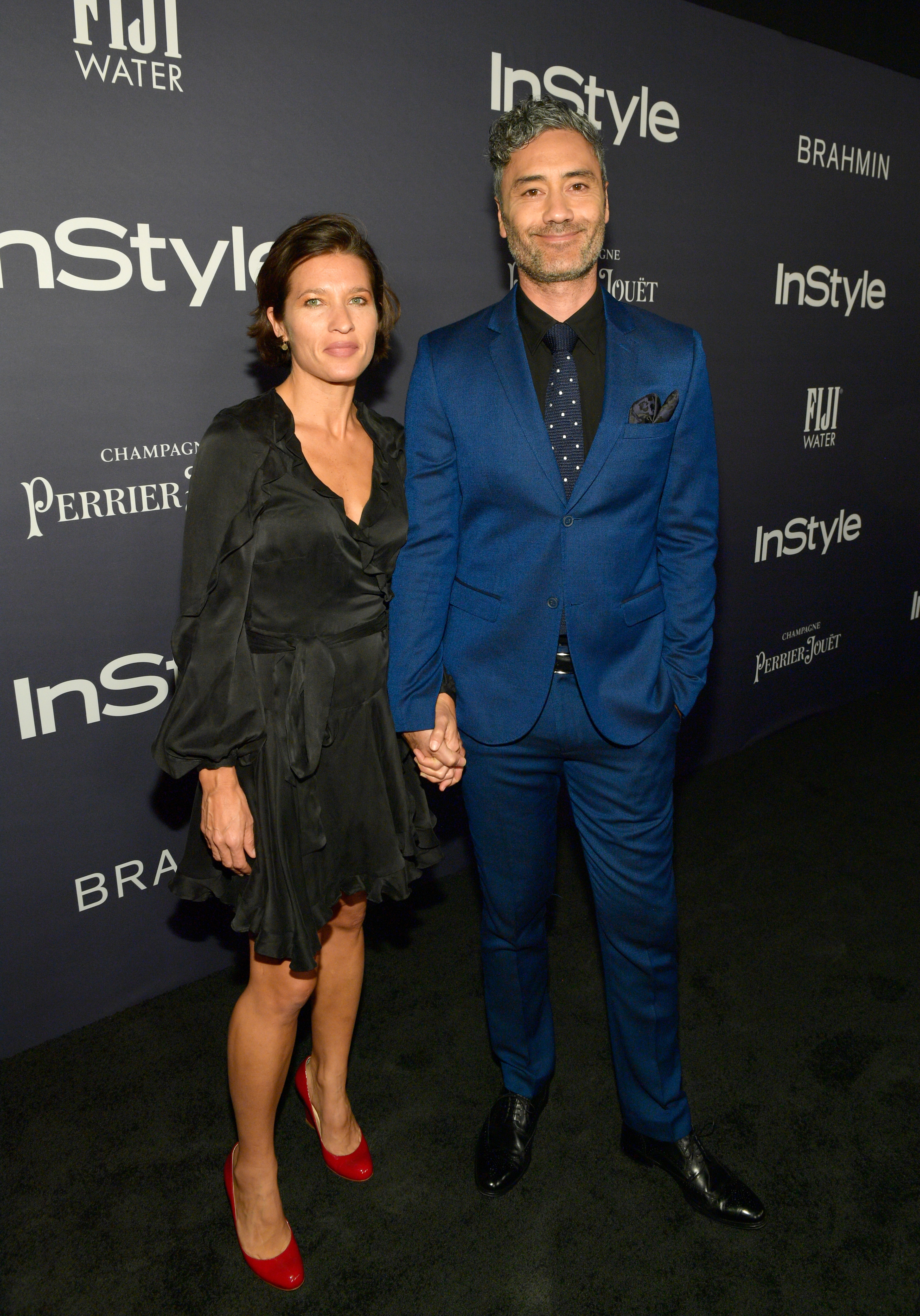 The former couple holding hands at a media event, with Chelsea in a dress and heels, and Taika in a tailored suit