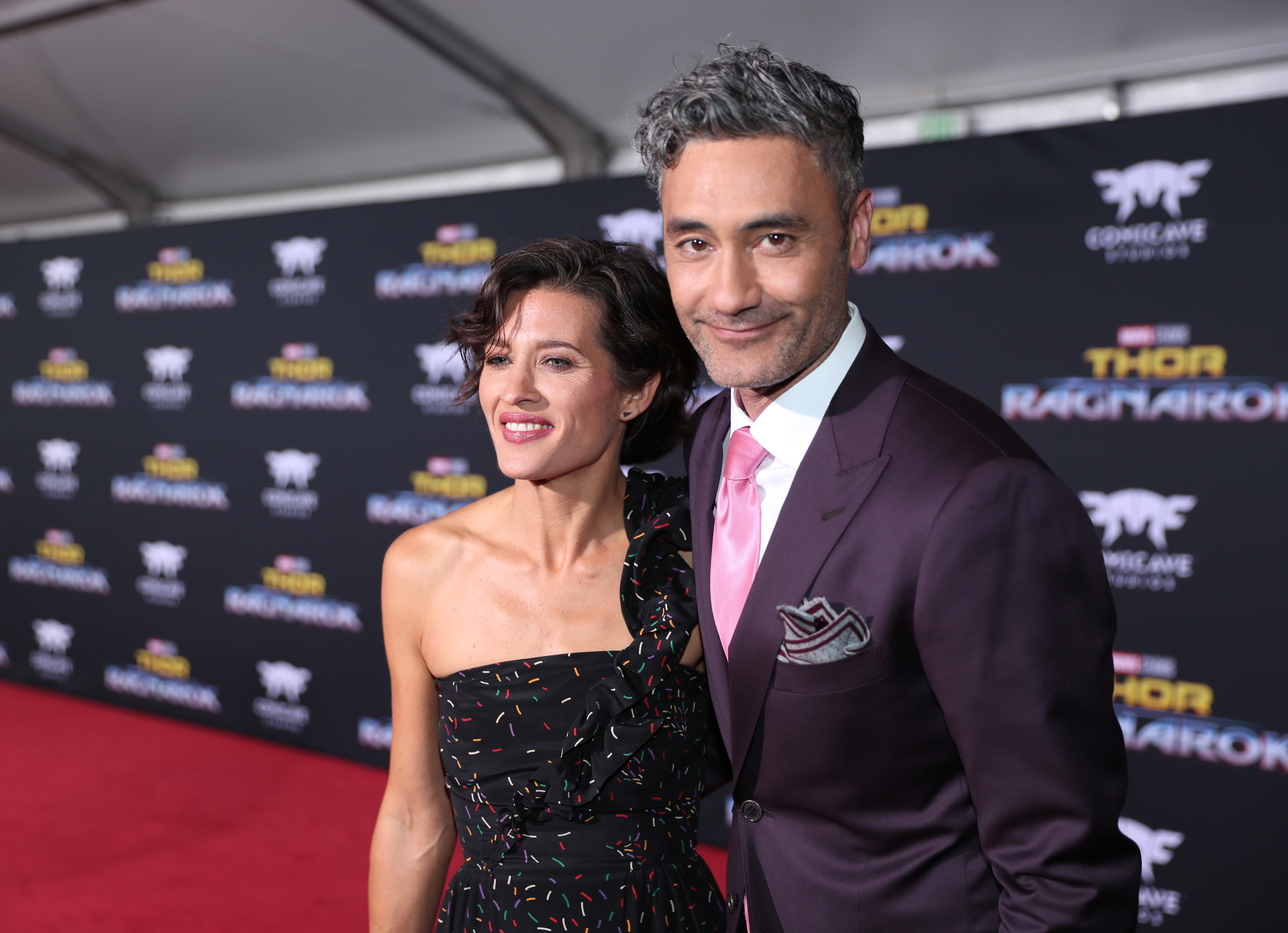 Chelsea and Taika posing on the red carpet, she in a patterned dress and he in a suit with a pink tie