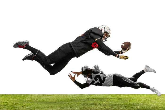 Two football players mid-air, one diving to catch the ball, the other reaching to block