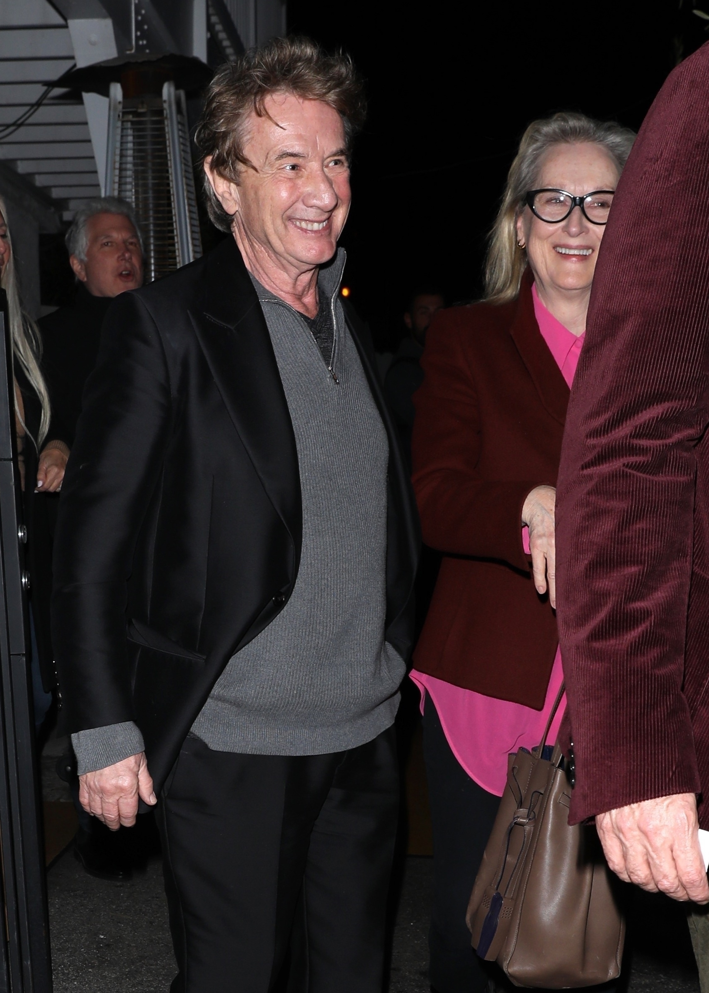 Martin Short in a black blazer smiles beside a woman in a maroon jacket on a night out