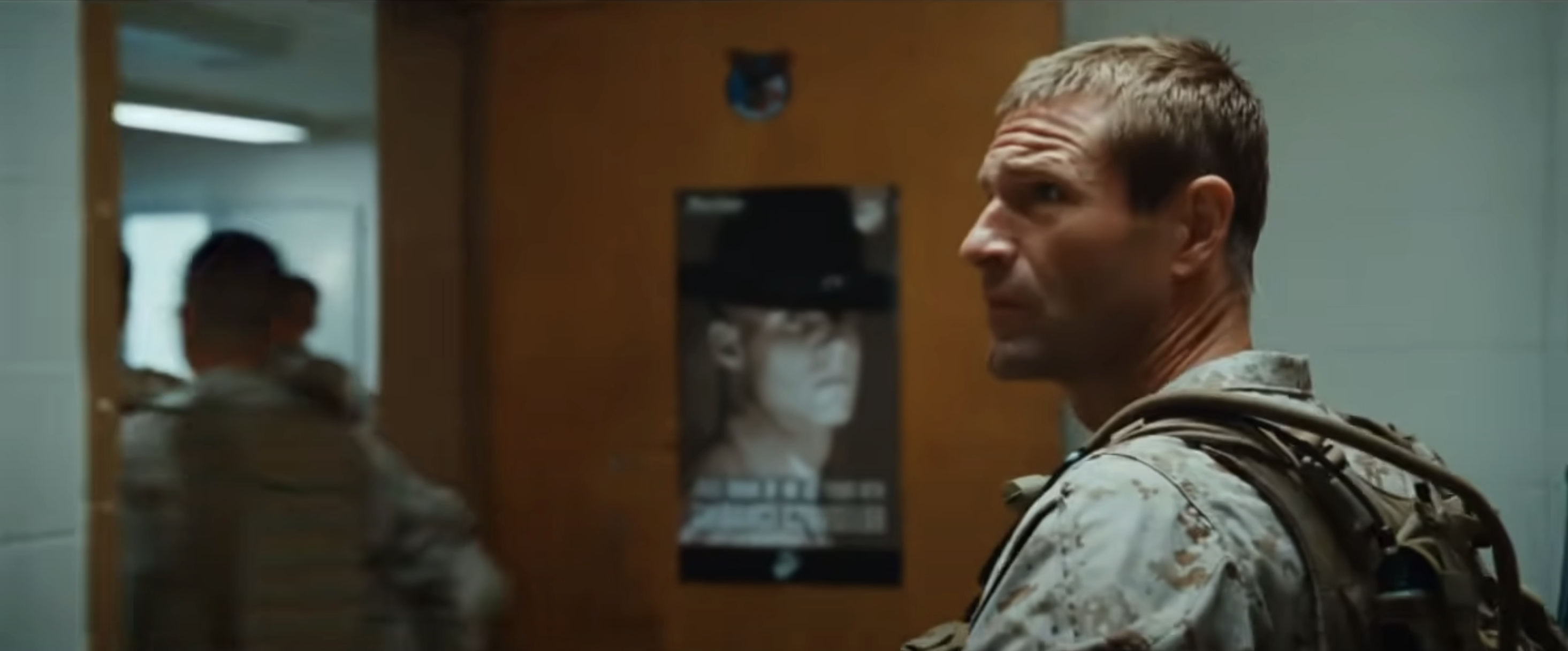 Michael in military gear standing in a hallway, looking back over his shoulder, next to a door with a poster