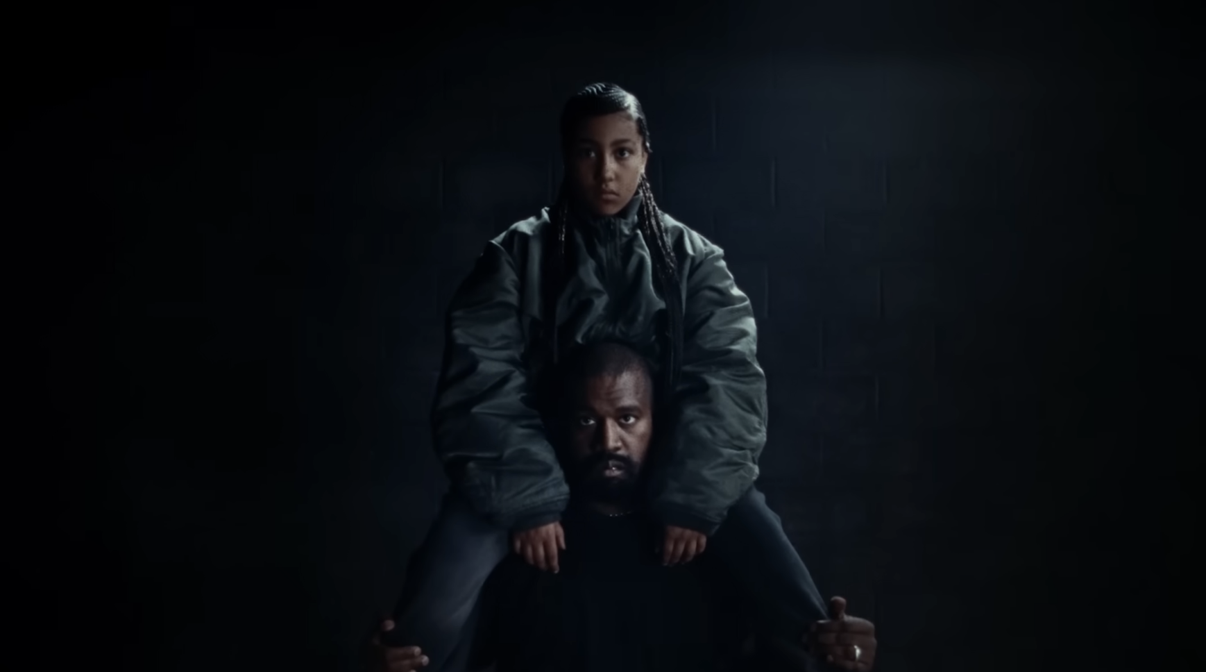Two individuals in oversized jackets against a dark background, one seated and one standing behind with arms resting on the seated person&#x27;s shoulders