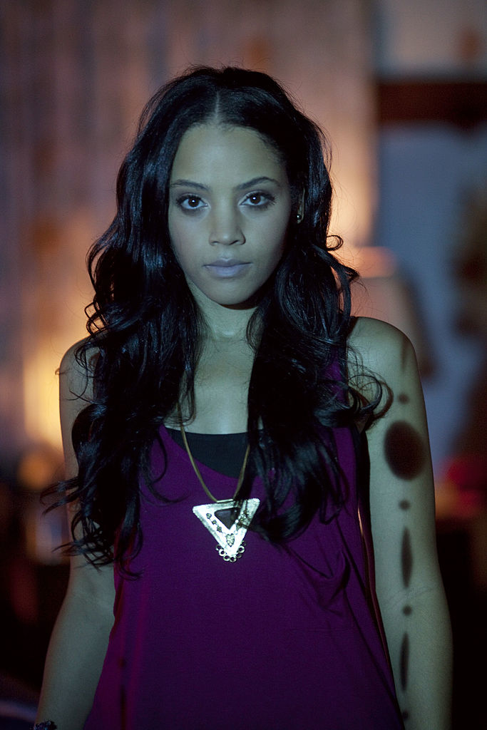 Bianca Lawson in a purple tank top with a triangle pendant, looking directly at the camera with a serious expression