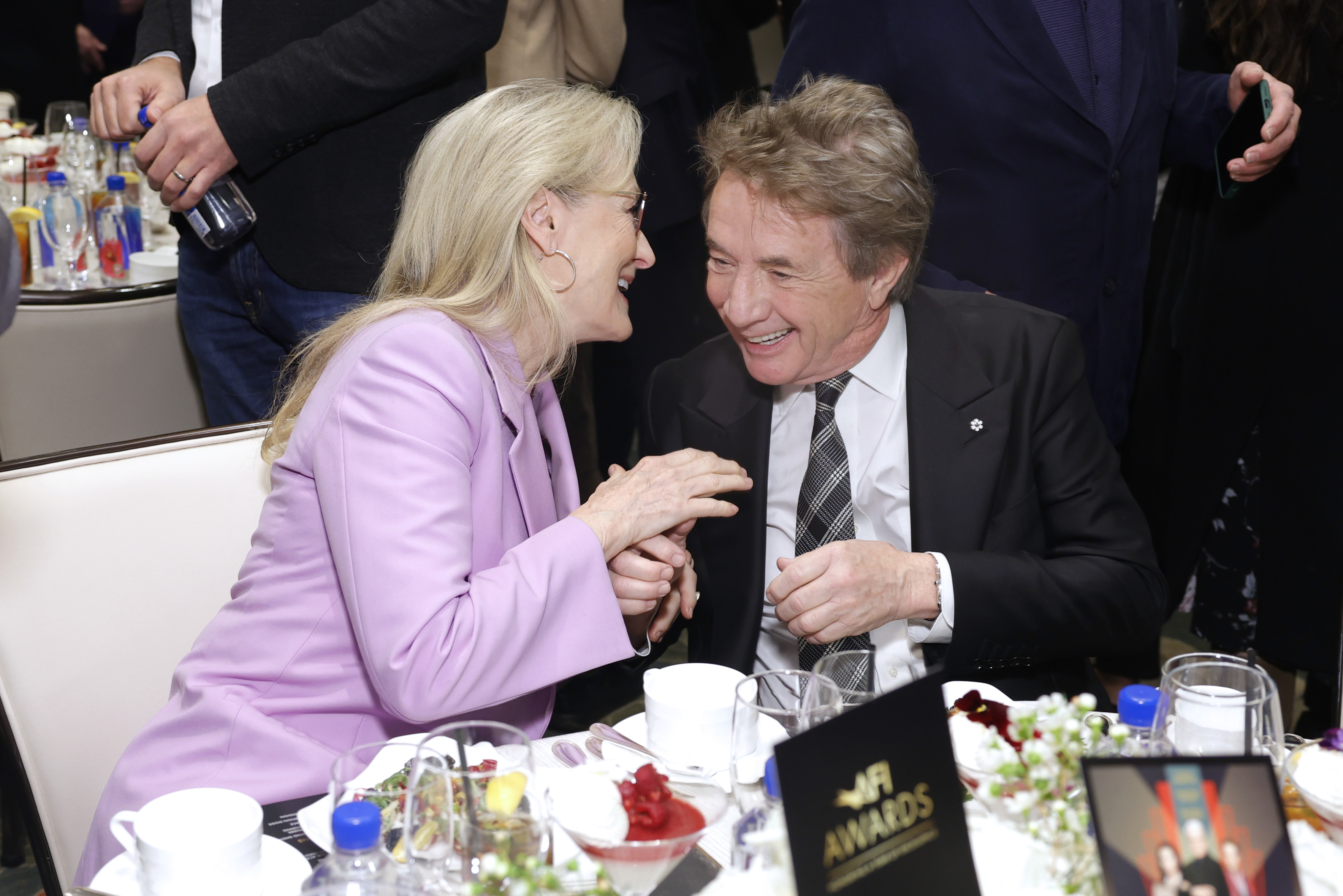 Two celebrities laughing together at a table during an event. Woman in a pink blazer, man in a suit with a striped tie