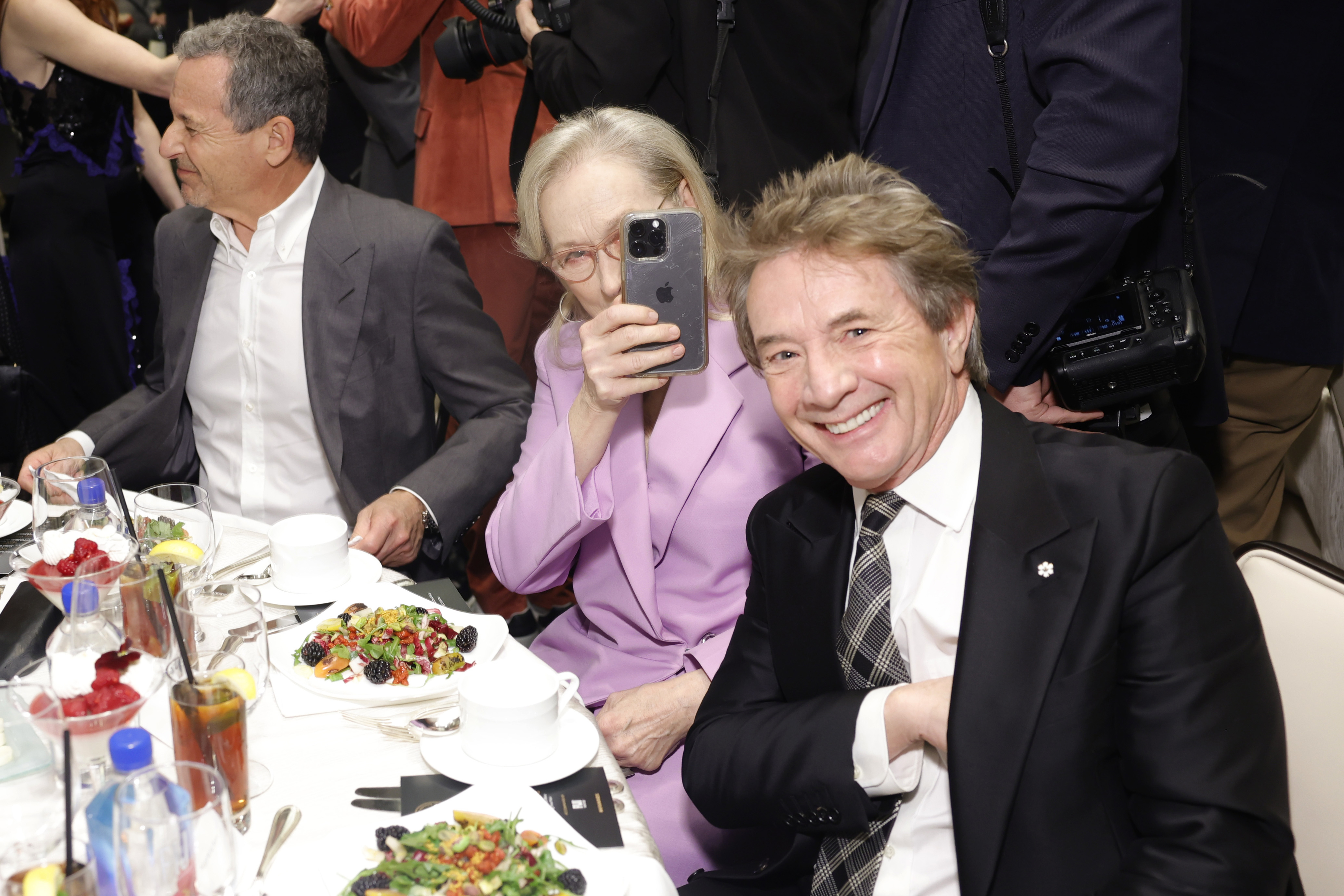 Two individuals at a table, one capturing a selfie, other smiling at the camera, amidst a social gathering