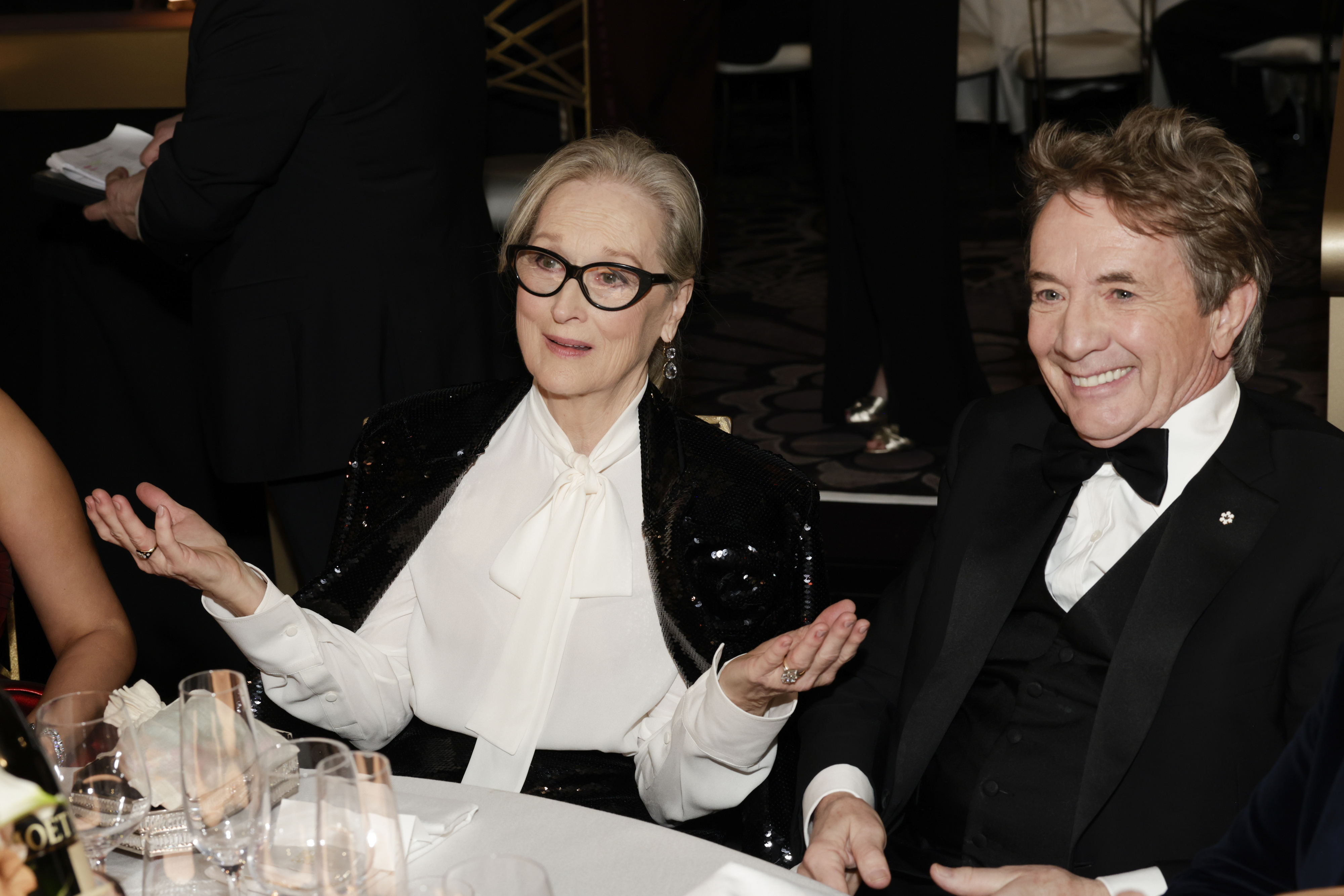Two celebrities seated at an event; woman in a white blouse with black details, man in a tuxedo