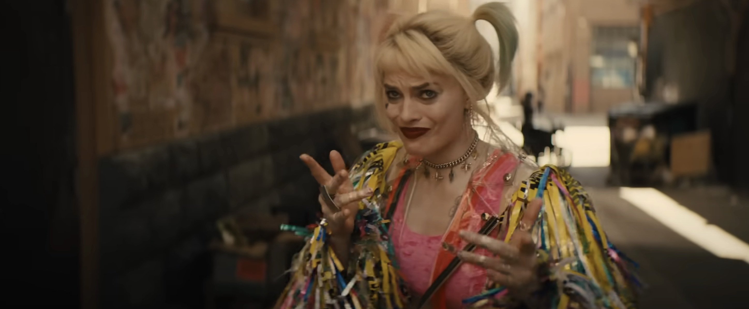 Harley Quinn smirks with a playful pose, wearing a colorful jacket with fringe details