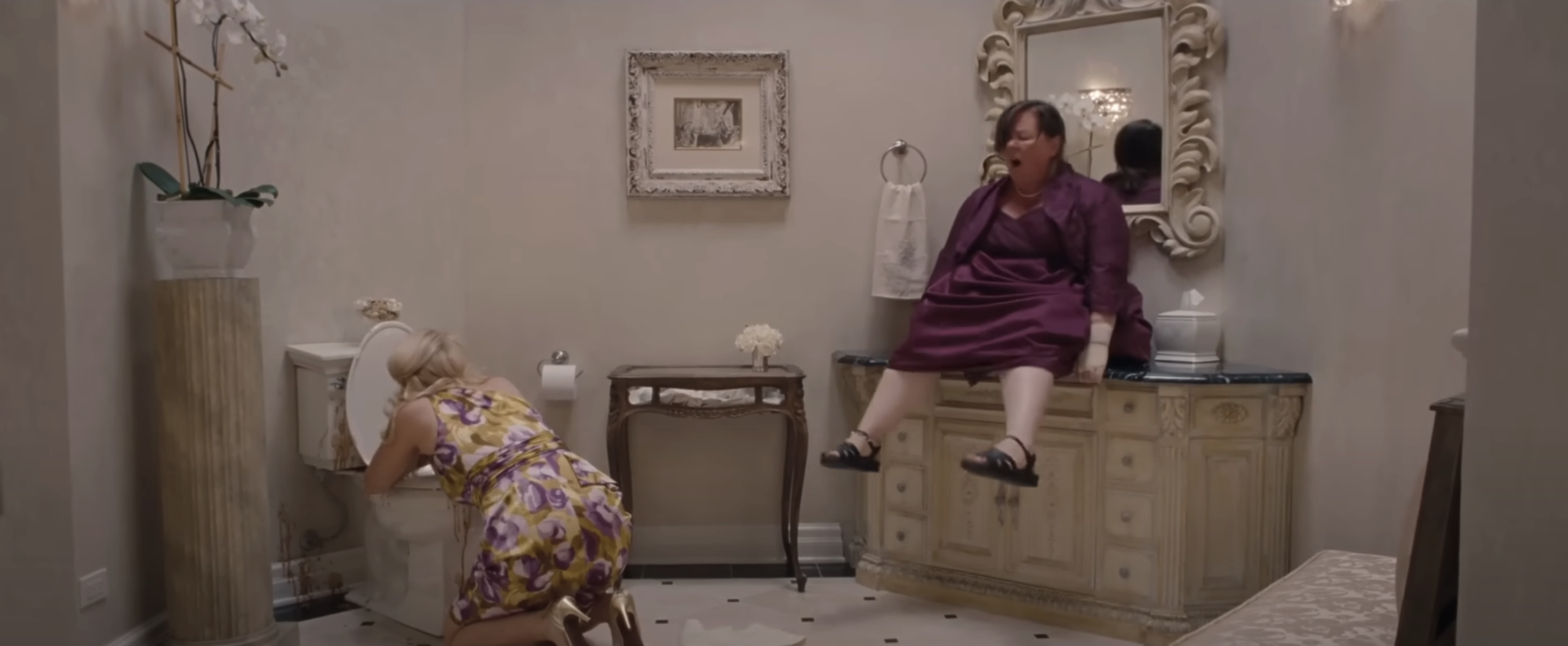 Two characters from Bridesmaids in a bathroom, one sitting on a sink, one kneeling by a toilet. Expressions suggest distress