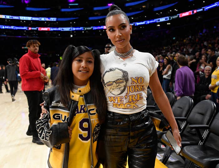 Two individuals pose at a basketball game, one wearing a Lakers jacket and the other in a graphic tee and shiny trousers