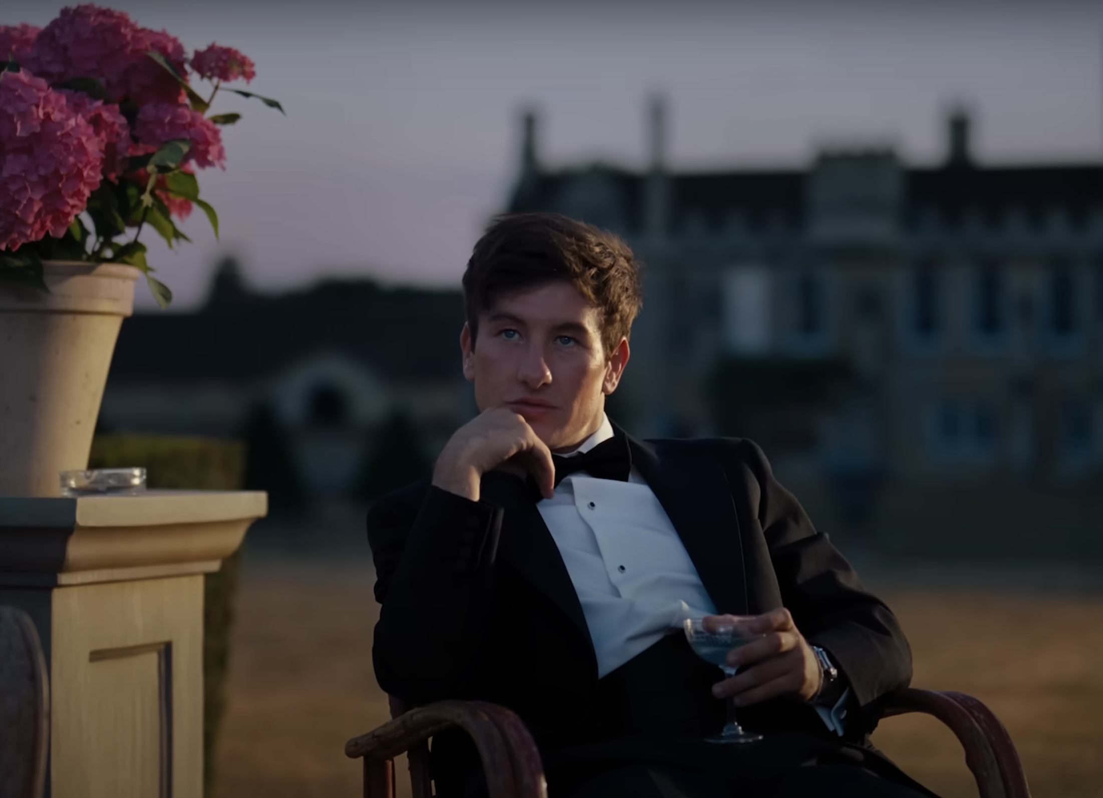 Oliver in a tuxedo sits contemplatively with a drink, outdoors at dusk, with a castle in the background