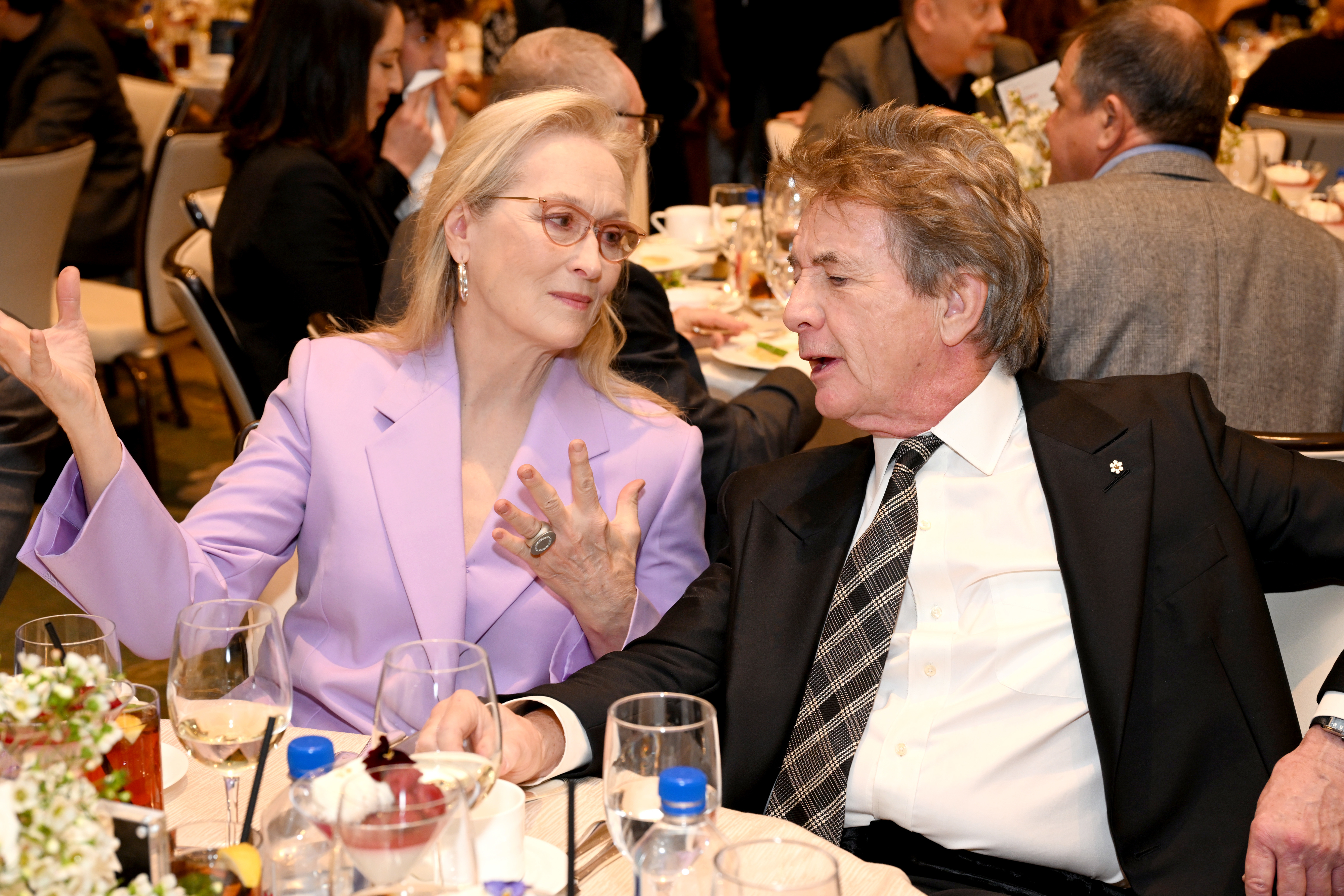 Two individuals sitting at a table engaged in conversation, man in black suit and woman in pastel blazer