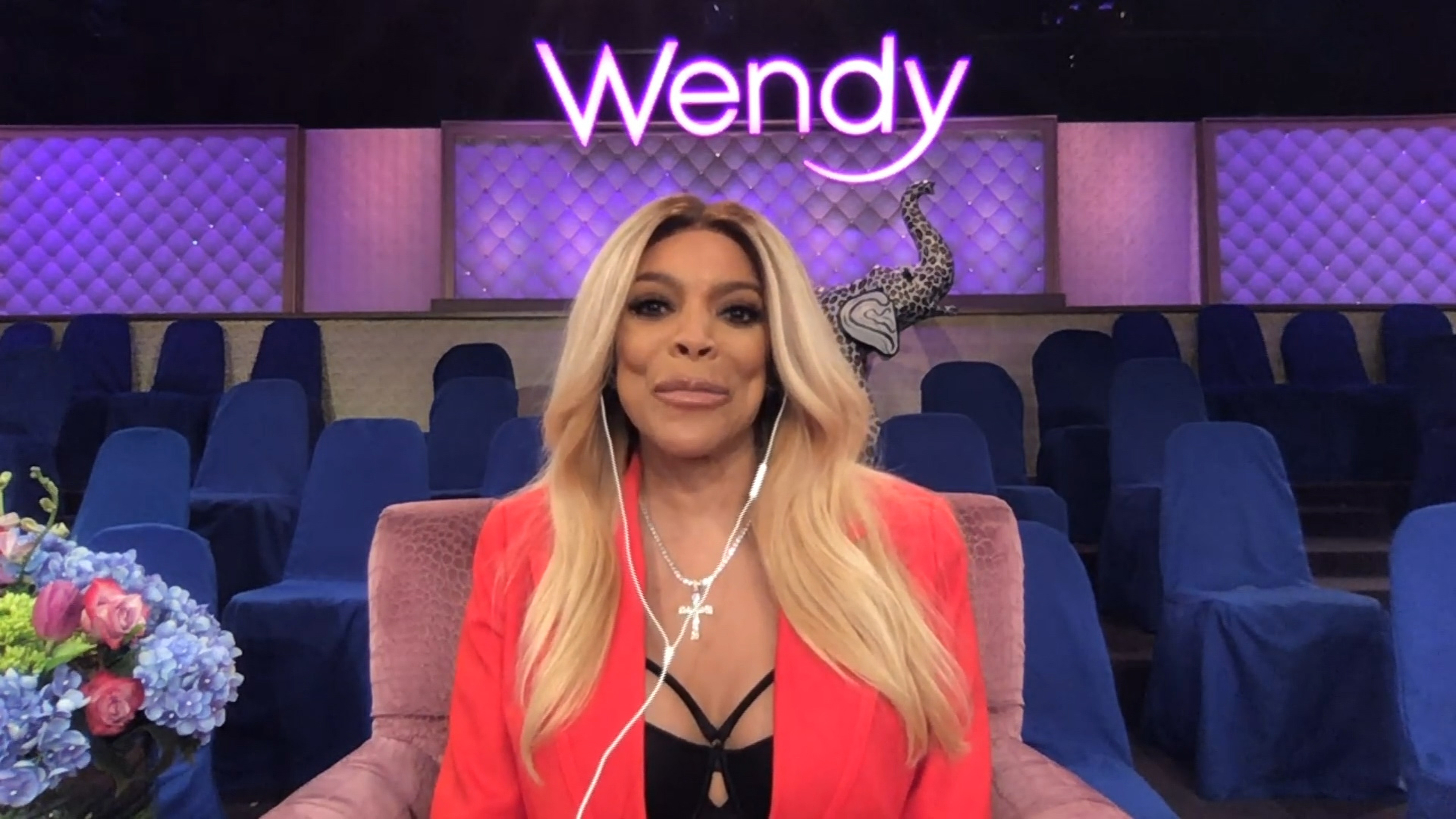 Wendy Williams seated in a studio, wearing a red blazer and black top, with a show logo in the background