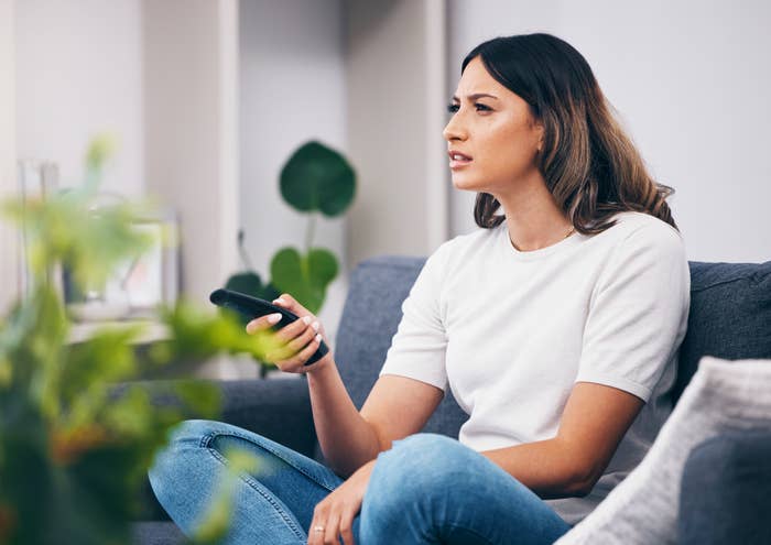 Woman sitting on a couch looking intently at a TV remote in her hand. She appears focused and confused