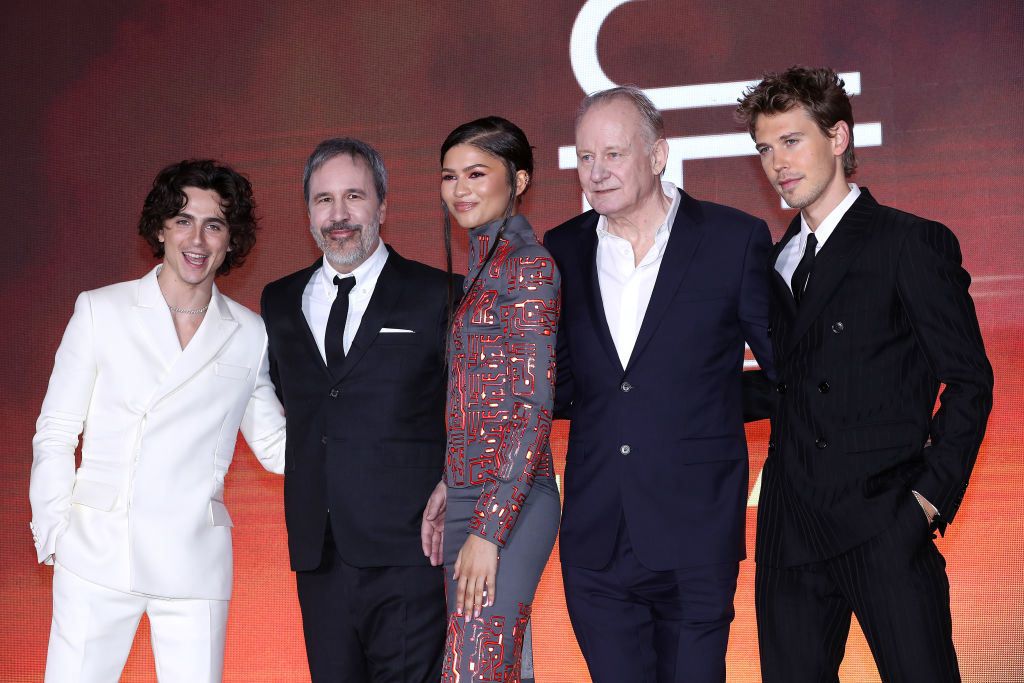 Five cast members posing at a film premiere; three men in suits, one woman in patterned attire, all smiling