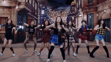 Grupo de seis integrantes de K-pop bailando en formación en un escenario con decoración urbana nocturna