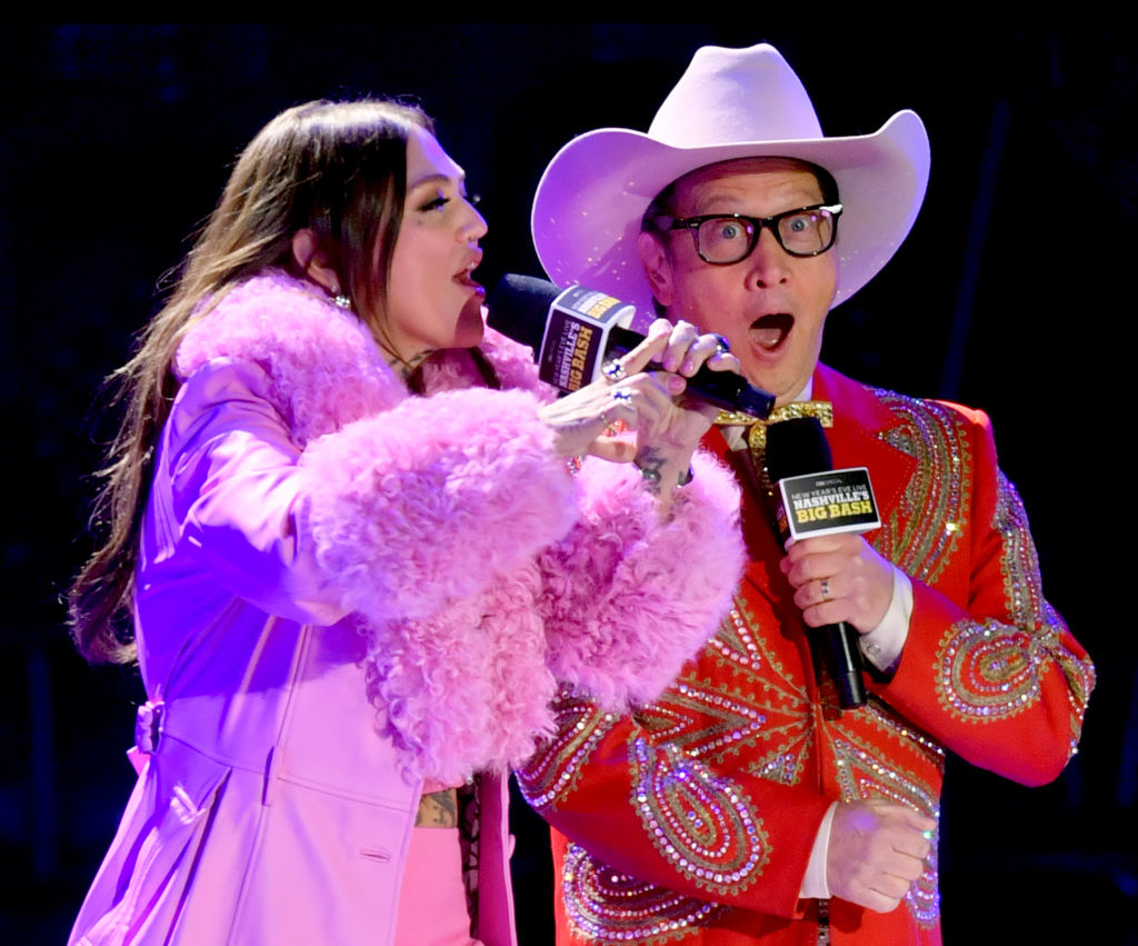 father and daughter on stage, Elle in a fluffy coat and Rob in a sequined outfit with a cowboy hat, singing into microphones