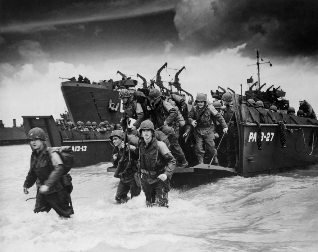 Soldiers disembark from a boat onto a shore, gear in hand, during an historical military operation