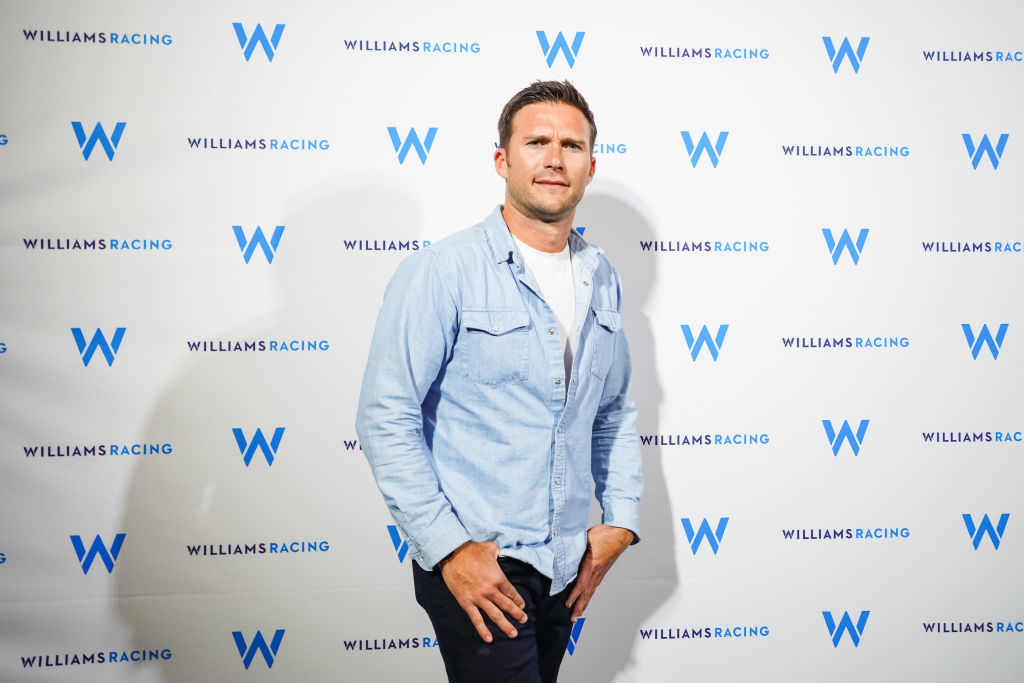 Scott in a denim jacket and shirt posing against a backdrop with Williams Racing logos