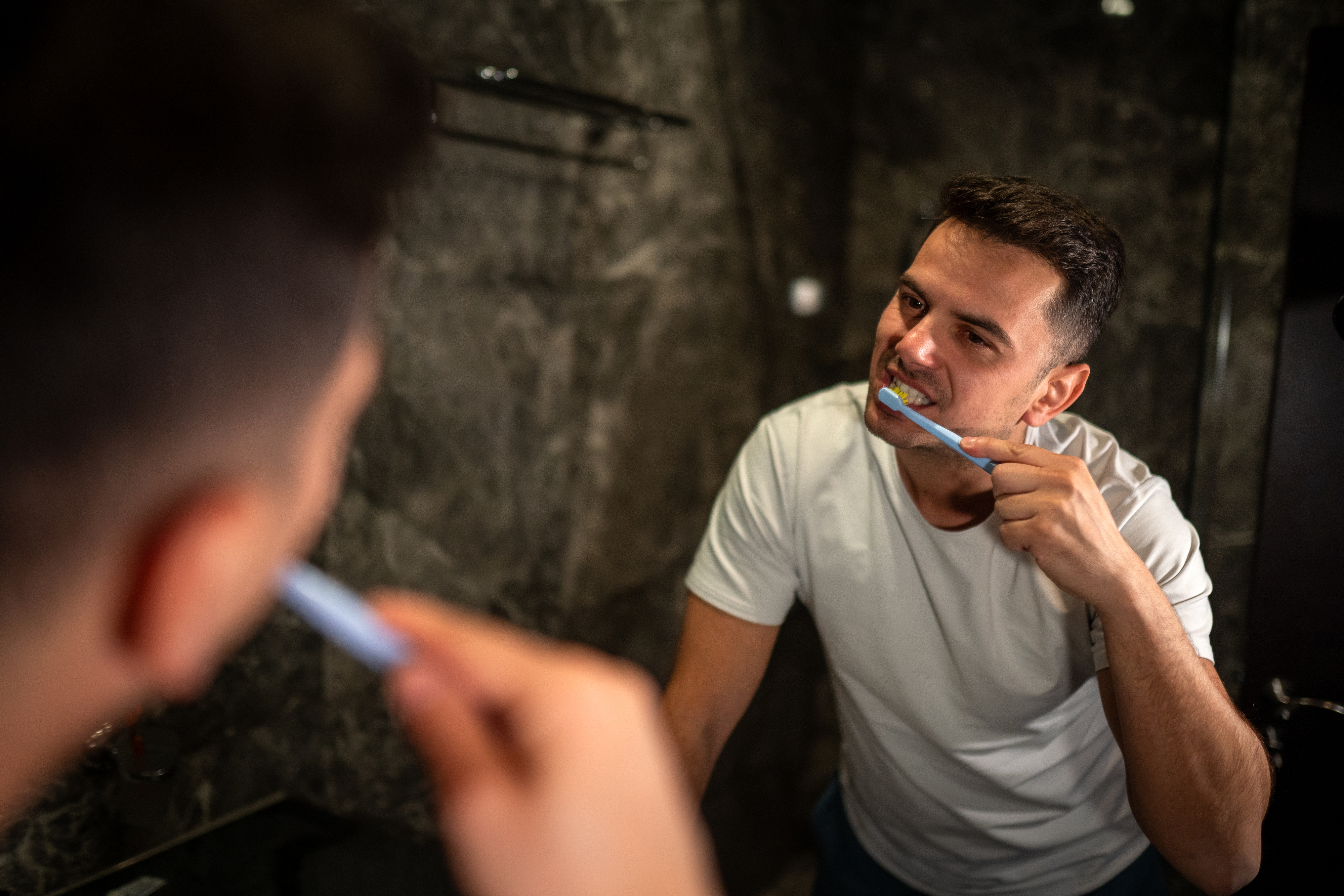 Man in a white shirt brushes his teeth reflected in a mirror in a bathroom setting