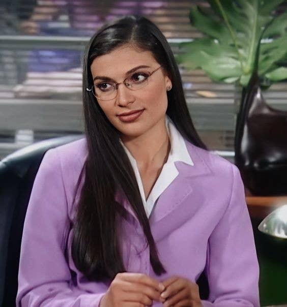 Mujer con traje sastre morado y lentes, sentada, posiblemente una actriz en set de TV