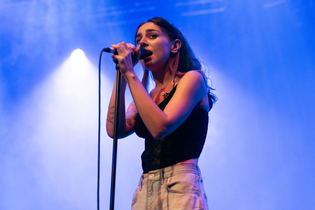 Samia with microphone on stage wearing a tank top and light pants, performing at a concert