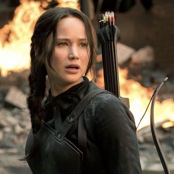 Katniss de Los Juegos del Hambre con arco y carcaj, expresión preocupada, fondos destrozados