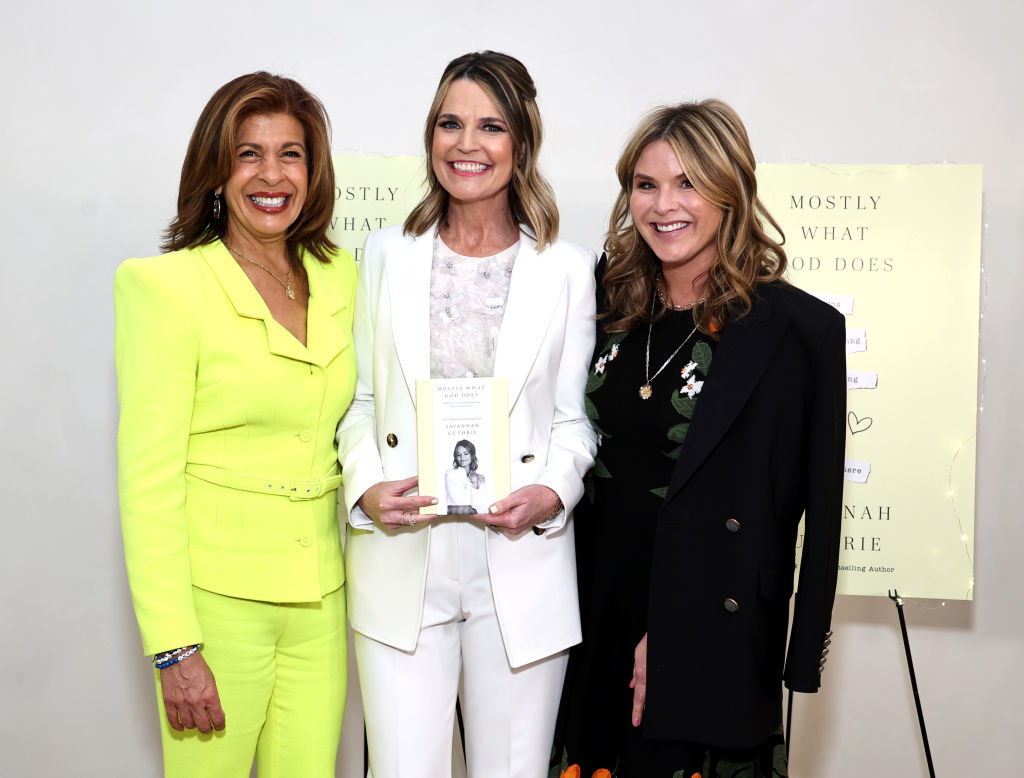 Three women, one in a yellow suit, another in white, and the third in black with floral accents, stand together holding a book