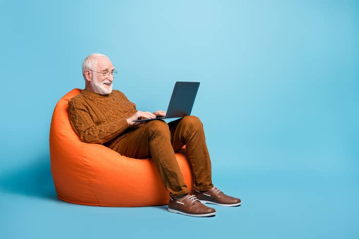 Elderly man smiling, seated on an orange bean bag and using a laptop