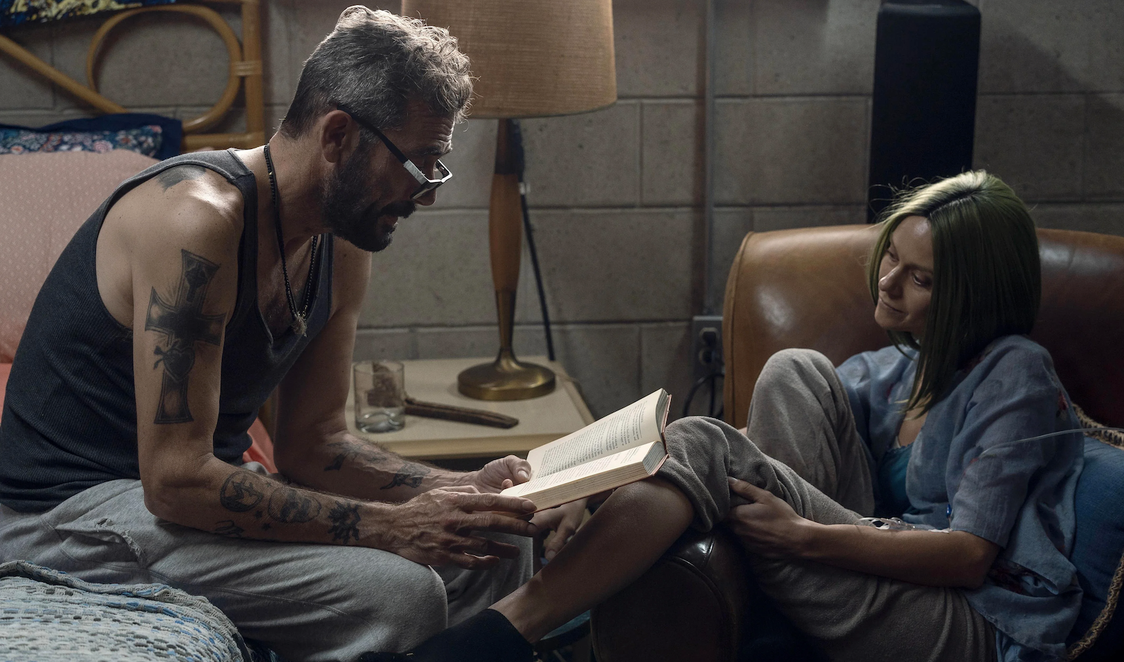 A man with tattoos and a tank top reads a book alongside a young girl in a casual setting
