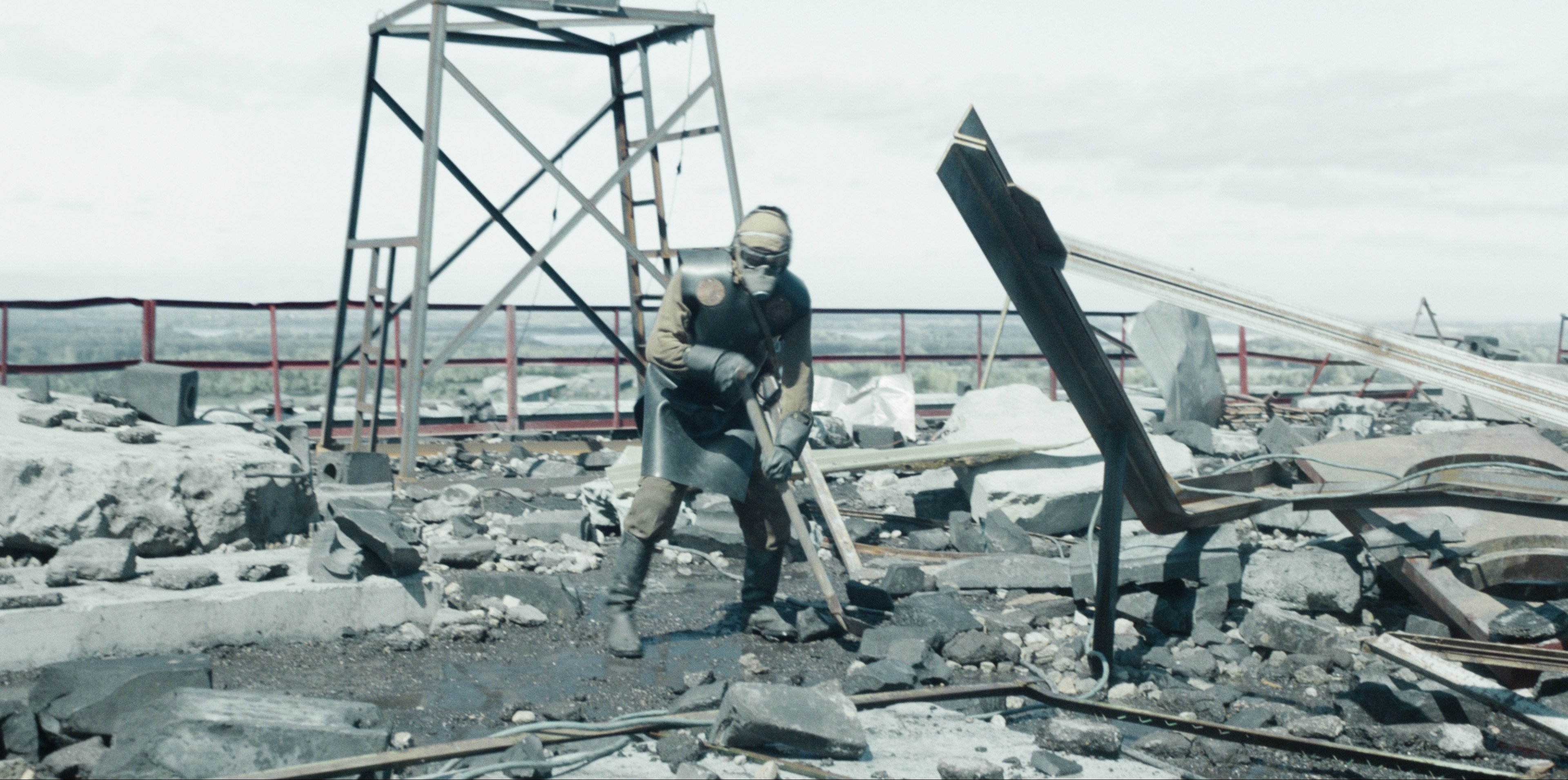 Person in protective gear kneels among industrial debris with a structure in the background