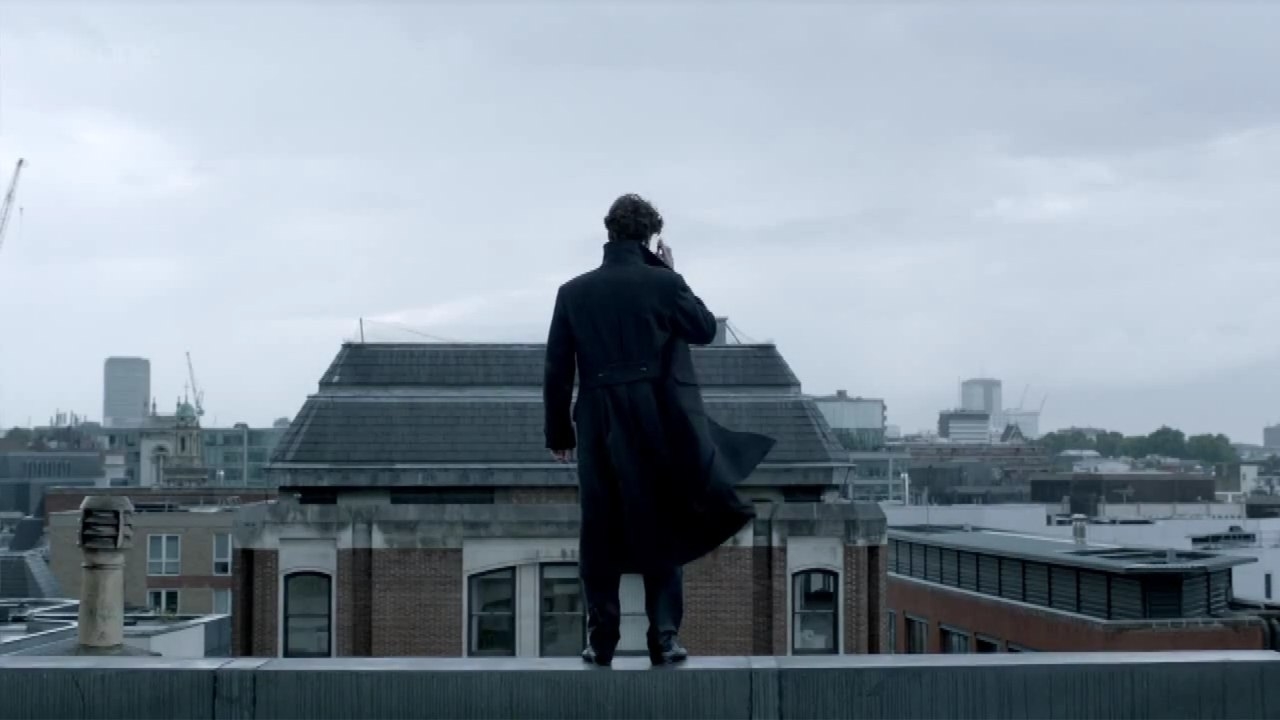 Sherlock Holmes stands on a rooftop overlooking a cityscape, dressed in his iconic coat