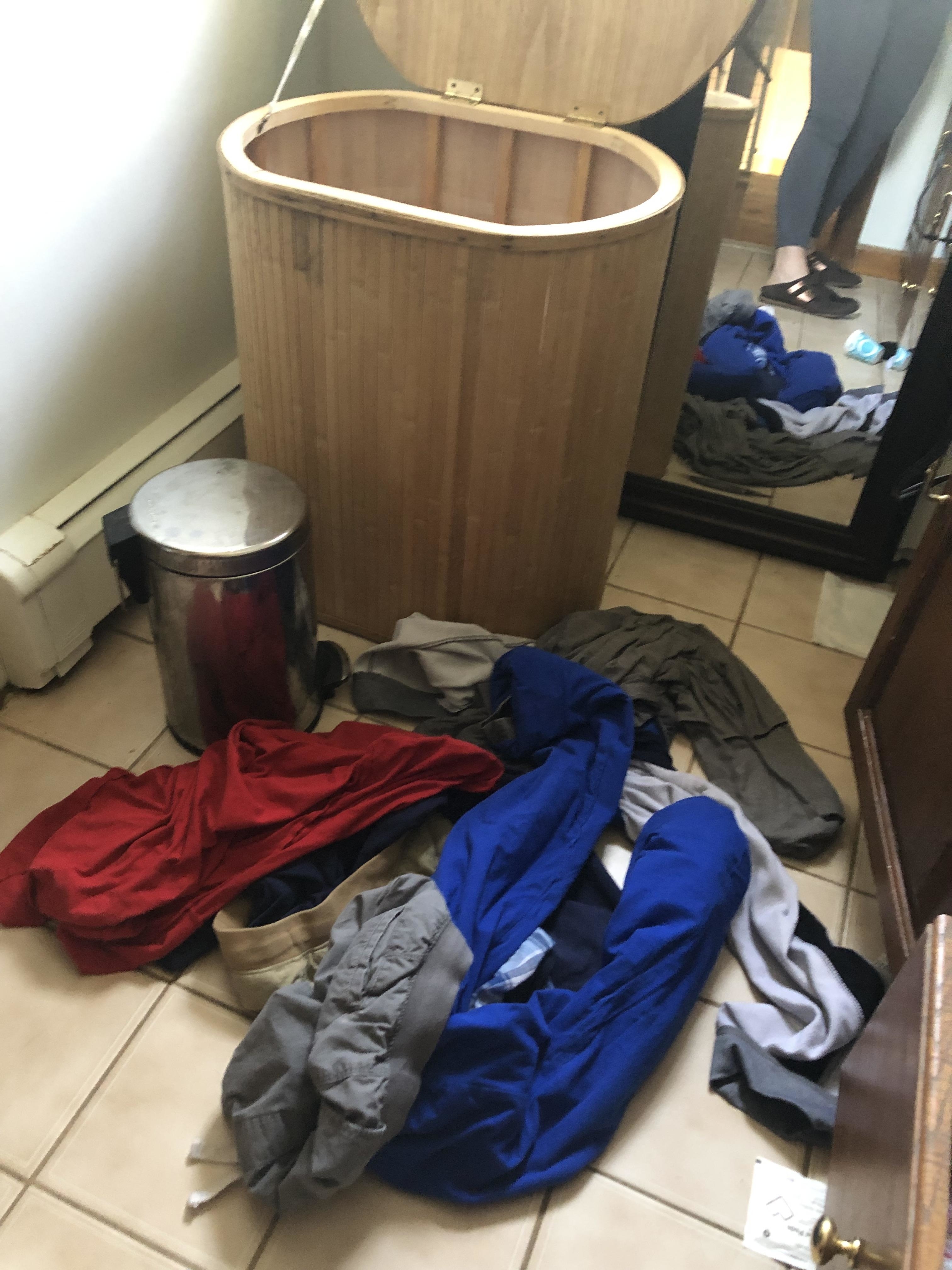 A cluttered room with clothes scattered on the floor and an open laundry hamper. Reflection of a person in the mirror